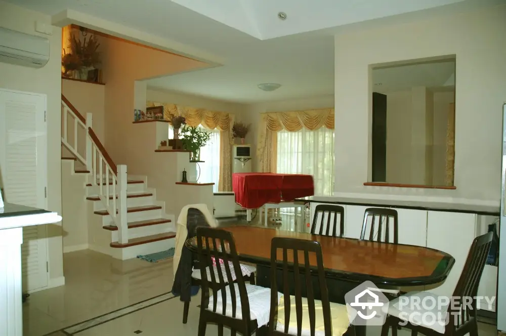 Spacious dining area with elegant wooden table set, adjacent to a cozy living space with a grand staircase, bathed in natural light from large windows.