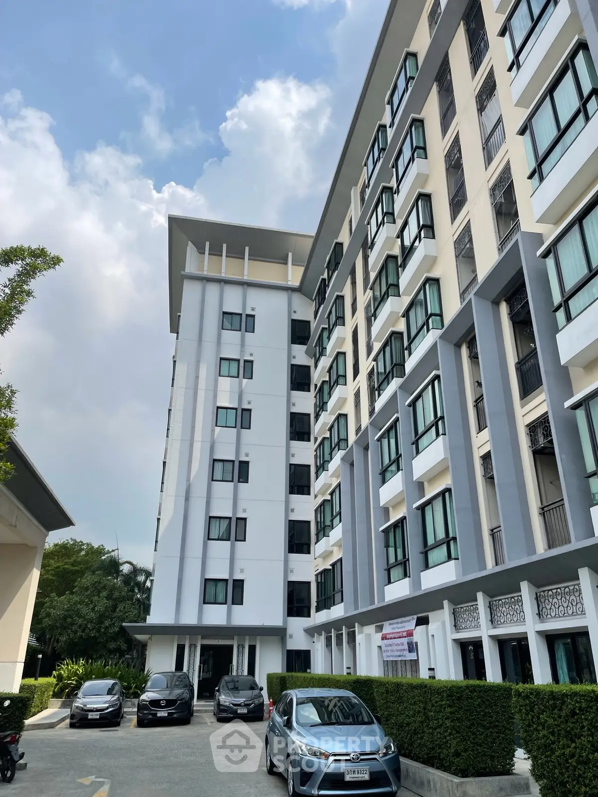 Modern multi-story residential building with parking area and lush greenery under a clear blue sky.