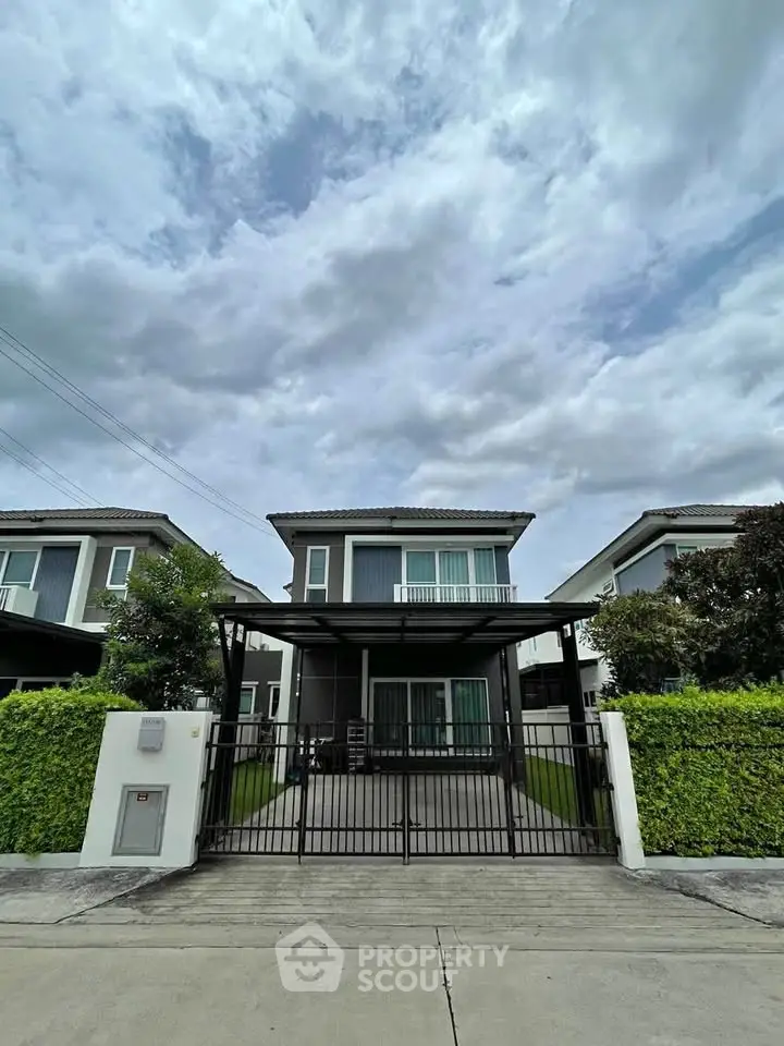 Modern two-story house with gated driveway and lush greenery under a cloudy sky.
