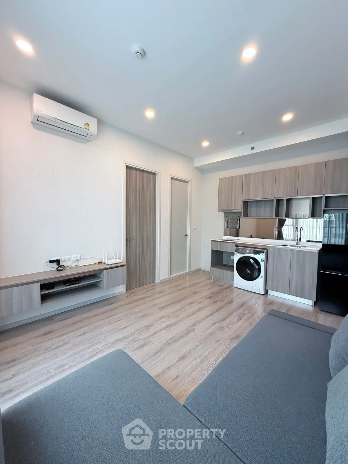 Modern living room with sleek kitchen and washing machine, featuring wooden flooring and ample lighting.