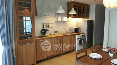 Modern kitchen with wooden cabinetry, washing machine, and dining area, featuring open layout and natural light.