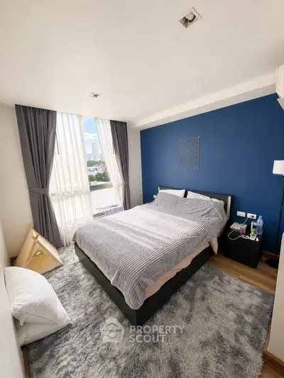 Modern bedroom with large window and city view, featuring cozy decor and stylish blue accent wall.
