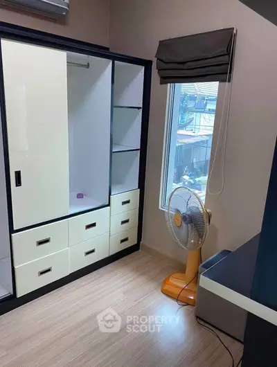 Cozy bedroom with built-in wardrobe and window view, featuring a standing fan and modern decor.