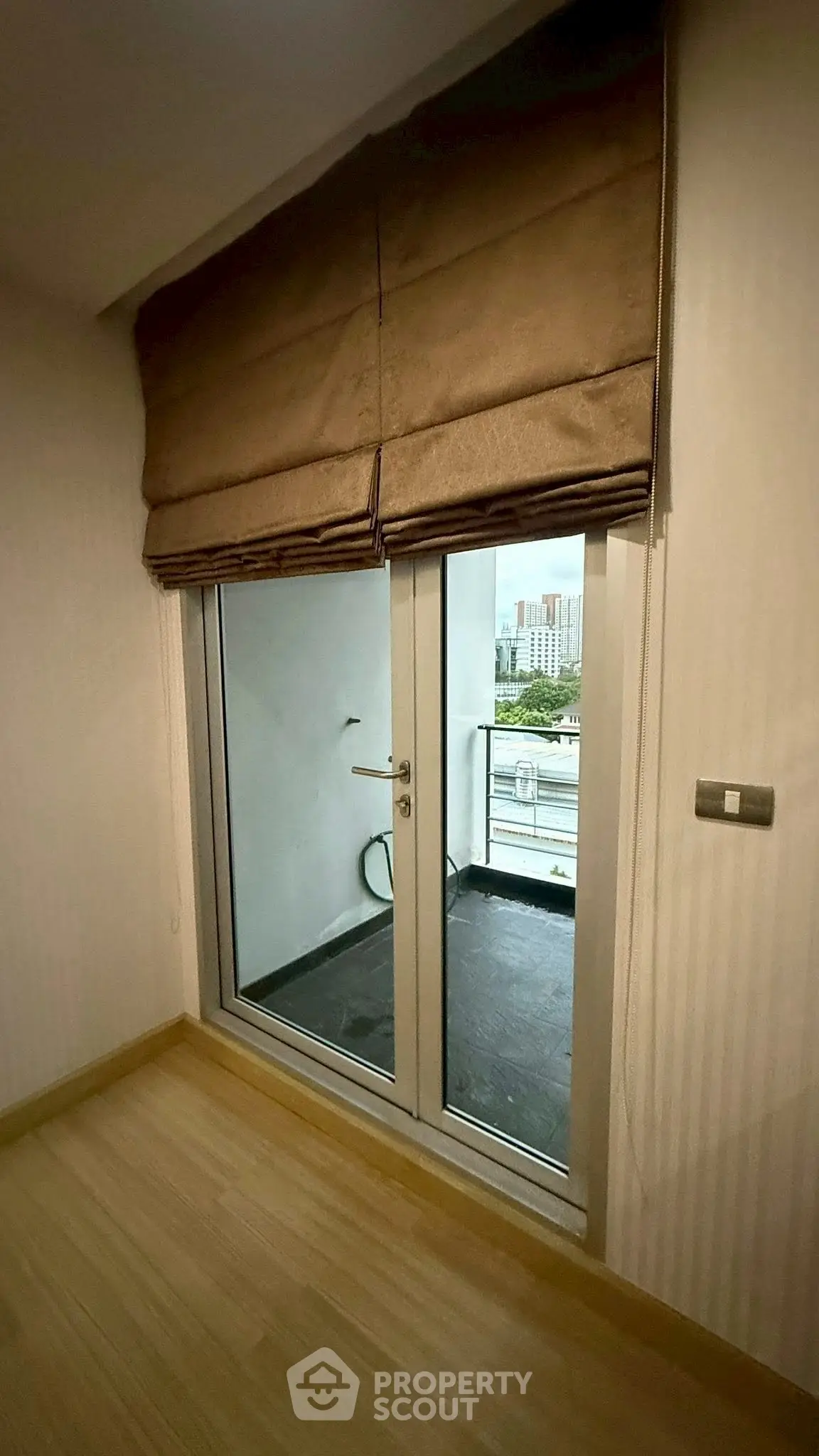 Cozy room with balcony access and city view, featuring elegant window blinds and wooden flooring.