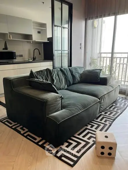 Modern living room with plush green sofa and open kitchen layout, featuring a stylish geometric rug and balcony view.