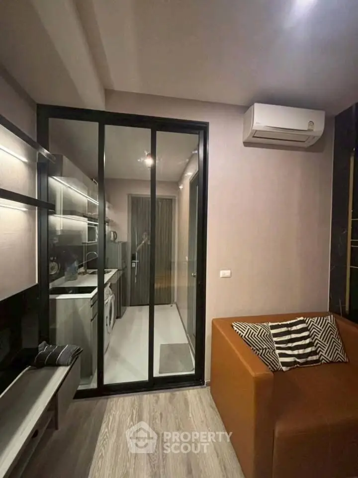 Modern living room with glass partition leading to sleek kitchen, featuring air conditioning and stylish decor.