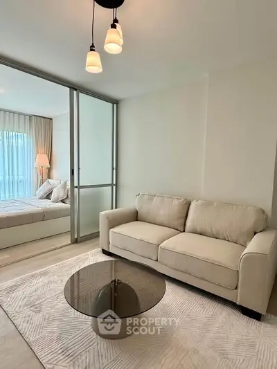 Modern living room with beige sofa and glass coffee table, adjacent to a cozy bedroom with natural light.
