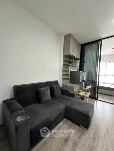 Modern living room with sleek gray sofa and open shelving, adjacent to a bedroom with glass sliding doors.