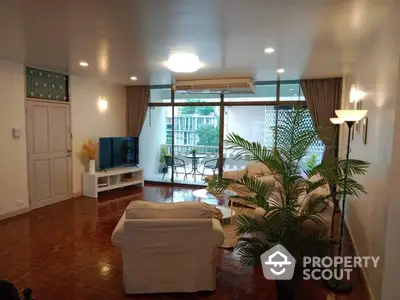 Spacious living room with polished hardwood floors, large windows draped in elegant curtains, and a cozy sitting area leading to a sunlit balcony with greenery.