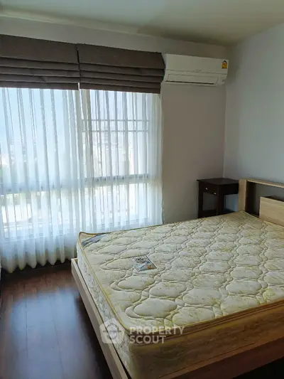 Bright bedroom with large window and air conditioning unit, featuring a comfortable bed and wooden flooring.
