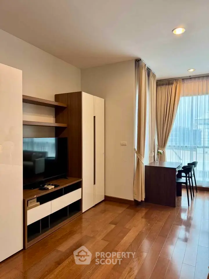 Modern living room with wooden flooring, TV unit, and large windows offering natural light.