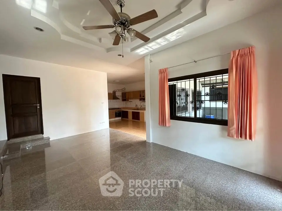Spacious living room with ceiling fan and natural light, adjacent to open kitchen.