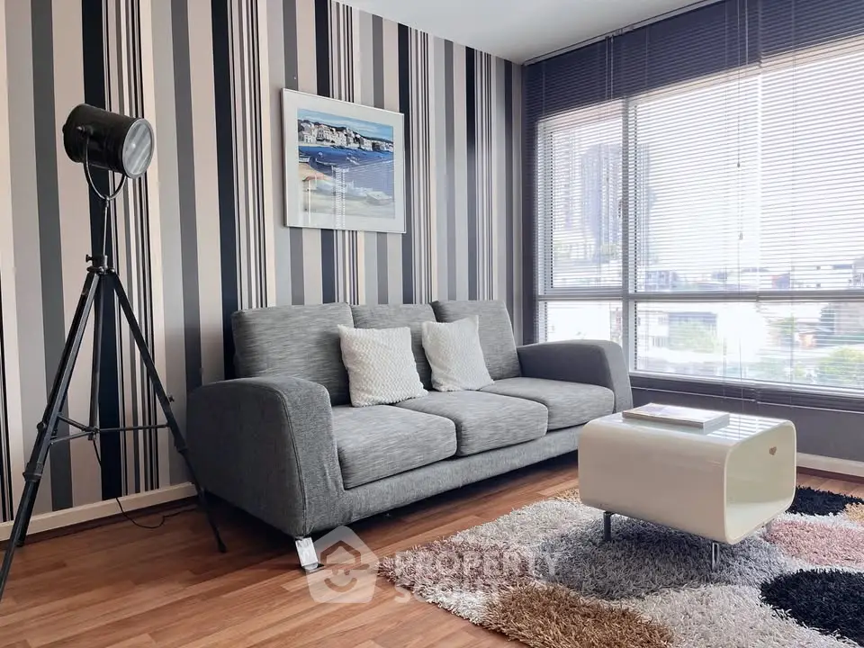 Stylish living room with modern decor, featuring a cozy gray sofa, striped wallpaper, and large window with city view.