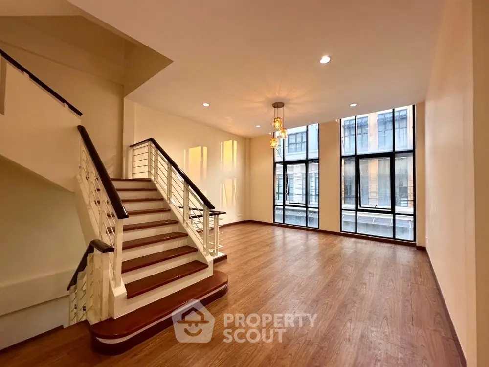 Spacious living room with elegant hardwood floors, large windows offering ample natural light, and a grand staircase leading to the upper level, perfect for luxurious urban living.
