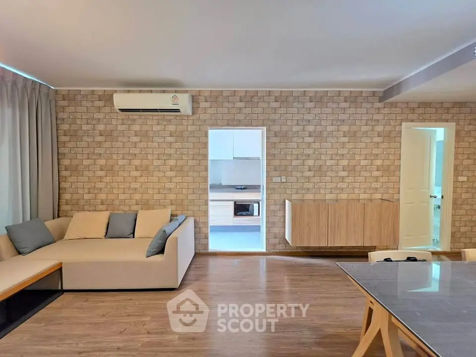 Spacious living room with modern decor and open kitchen layout, featuring a cozy sofa and stylish brick wall.