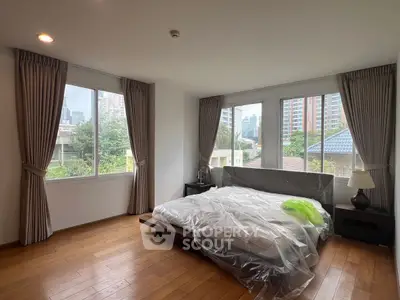 Spacious bedroom with large windows and city view, featuring elegant curtains and wooden flooring.