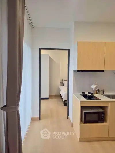 Modern apartment interior with compact kitchen and view into cozy bedroom, featuring sleek cabinetry and natural light.