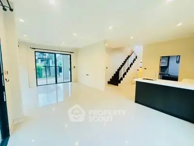 Spacious open-plan living room with modern kitchen and staircase, featuring large windows and bright lighting.