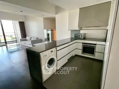Modern open-plan kitchen with washing machine and oven, leading to a bright living area with large windows.