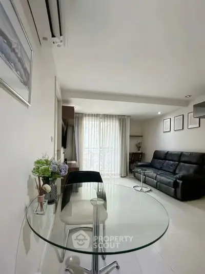 Modern living room with glass table and black leather sofa, bright natural light from large window.