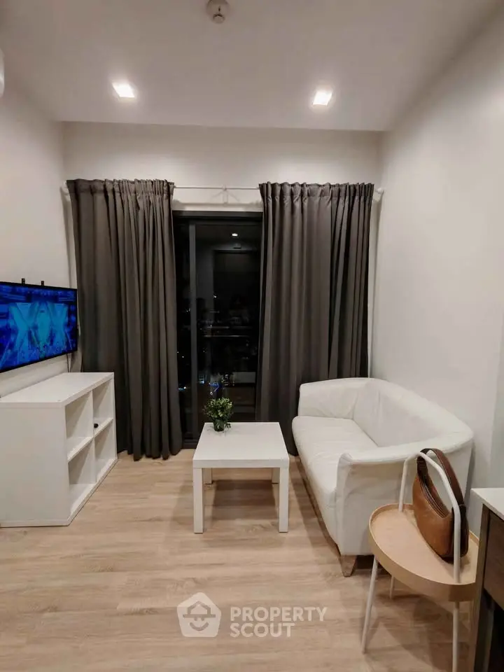 Cozy modern living room with white furniture and wooden flooring, featuring a TV and large window with curtains.