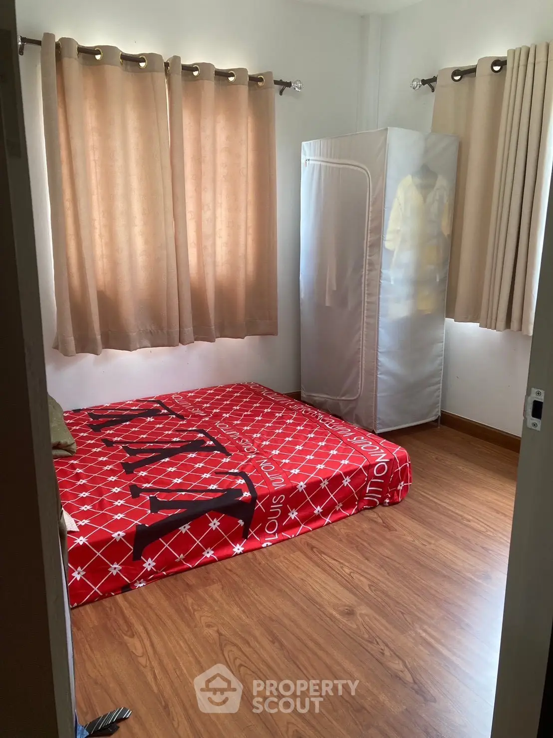Cozy bedroom with red bedding and wooden flooring, featuring natural light from curtained windows.