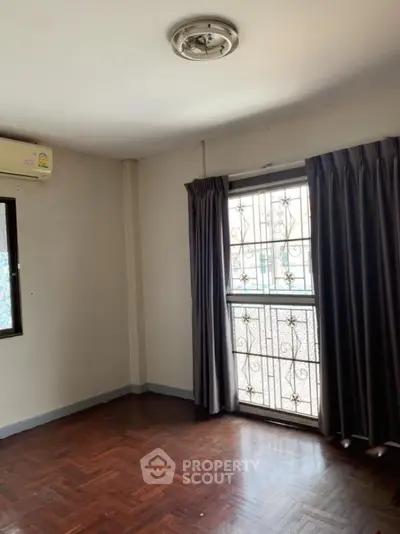 Spacious room with parquet flooring and large window in a well-lit apartment.