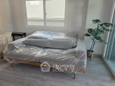Modern living room with new sofa wrapped in plastic, large window, and potted plant.