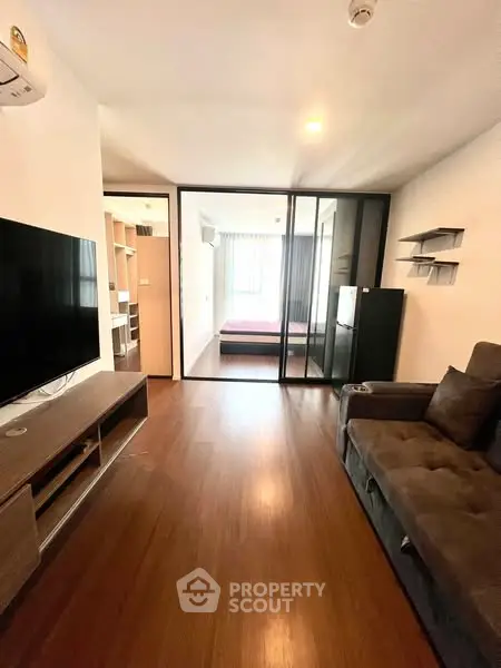 Modern living room with wooden flooring, large TV, and cozy sofa leading to a bedroom.