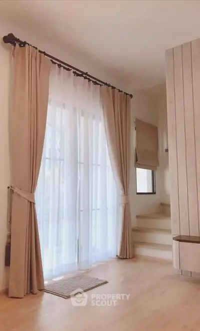Elegant living room with large windows and soft curtains, featuring a cozy staircase corner.