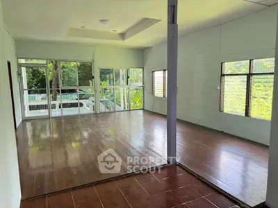 Spacious living room with wooden floors and large windows offering lush green views.