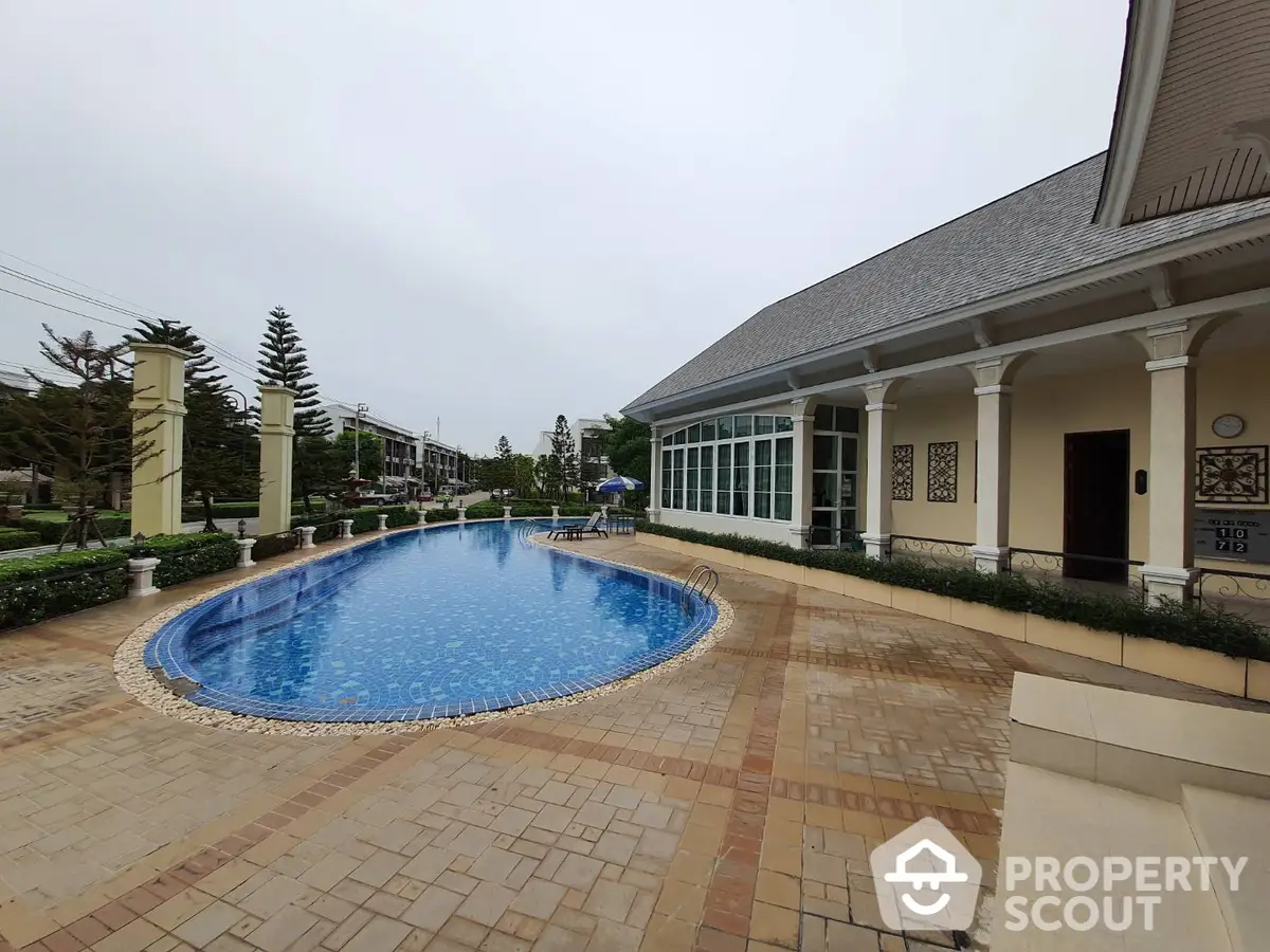 Elegant estate with a stunning blue pool surrounded by a beautifully tiled patio and manicured gardens, adjacent to a grand clubhouse.