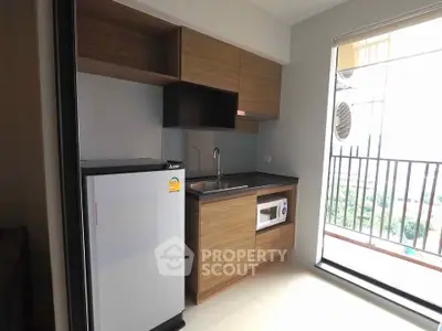 Modern compact kitchen with sleek wooden cabinets and appliances, featuring a fridge and microwave, next to a balcony.