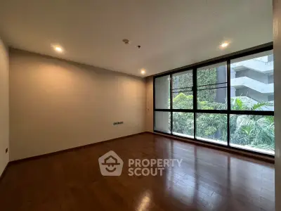 Spacious empty room with large windows and wooden flooring, offering a serene garden view.