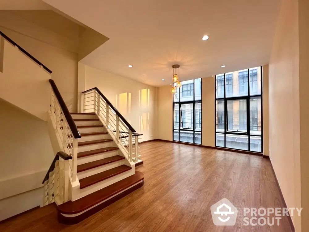 Spacious living room with elegant hardwood floors, large windows offering ample natural light, and a grand staircase leading to the upper level, perfect for luxurious urban living.