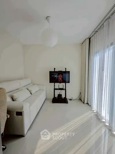 Bright living room with modern white sofa and wall-mounted TV, featuring floor-to-ceiling windows and sheer curtains.