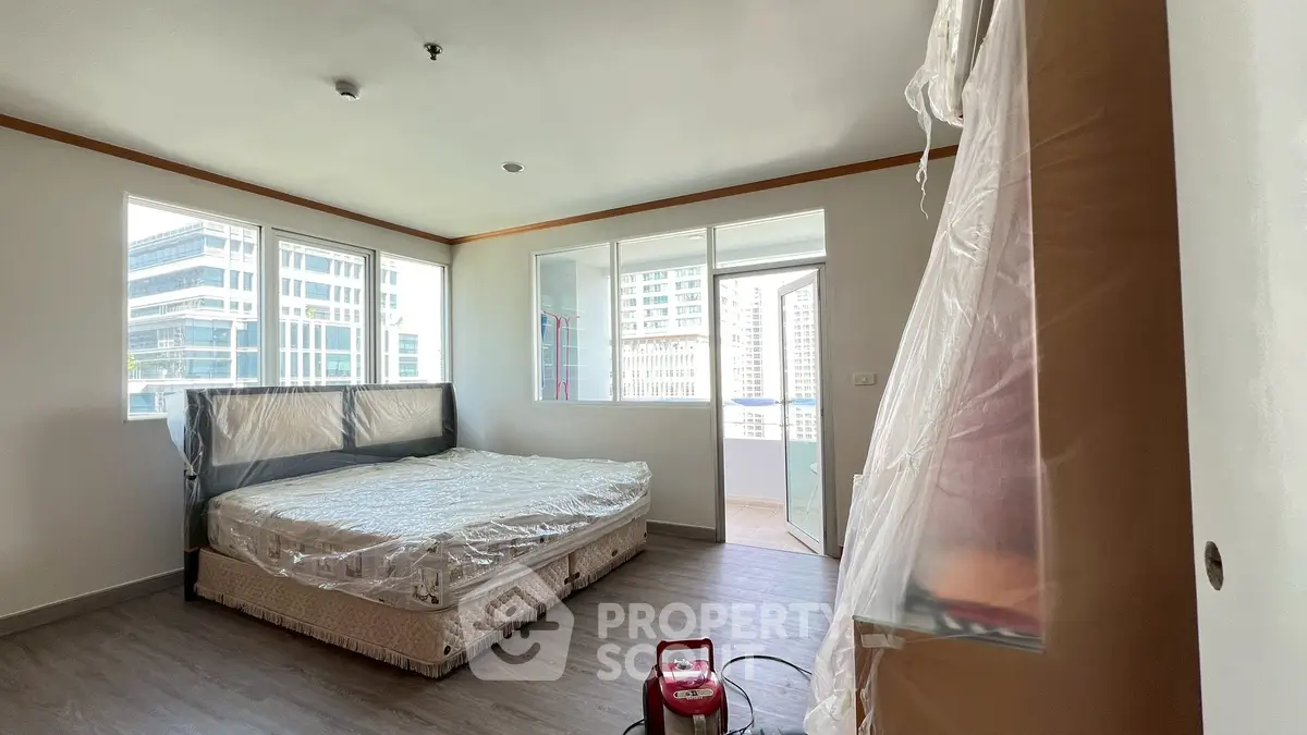 Spacious bedroom with large windows and city view, featuring a covered mattress and balcony access.