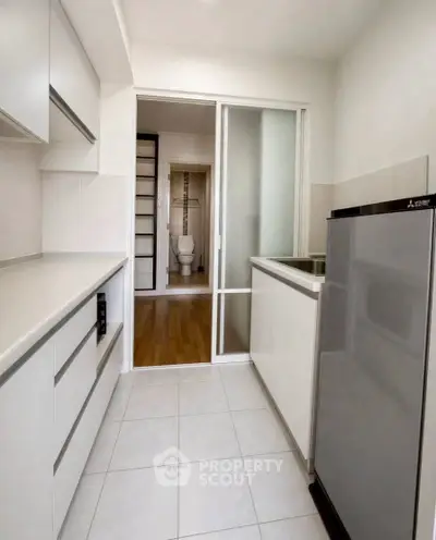 Modern kitchen with sleek cabinetry and compact fridge, leading to a bathroom with sliding door.