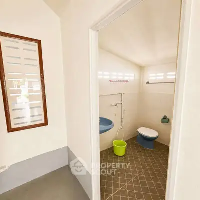 Minimalist bathroom with unique blue toilet and green bucket, featuring tiled floor and natural light.