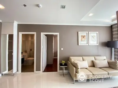 Spacious living room with elegant beige sofa, tiled flooring, and neutral color palette, leading to a well-lit bathroom and bedroom.