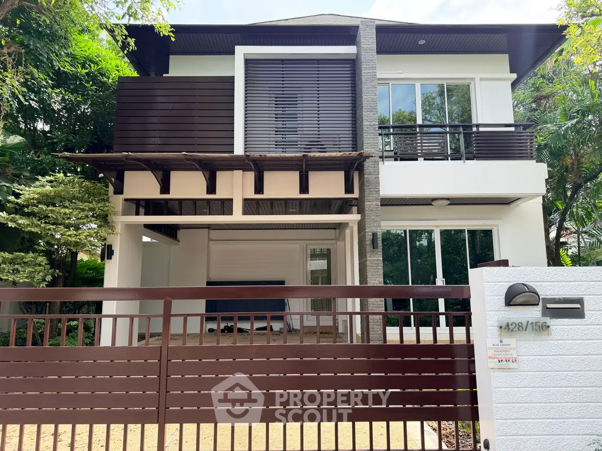 Modern two-story house with sleek design and lush greenery, featuring a gated entrance and large windows.