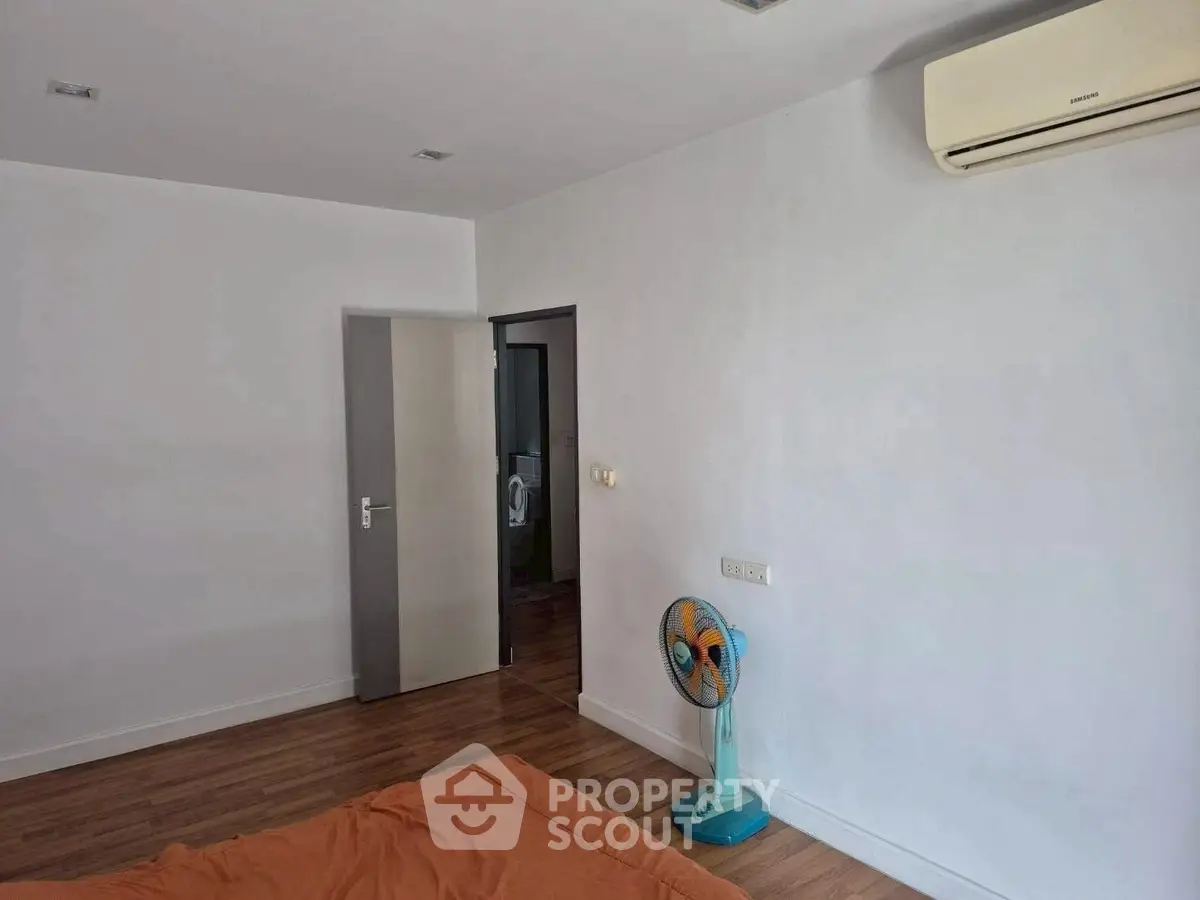 Minimalist bedroom with air conditioning and wooden flooring, featuring a simple fan and orange bedding.