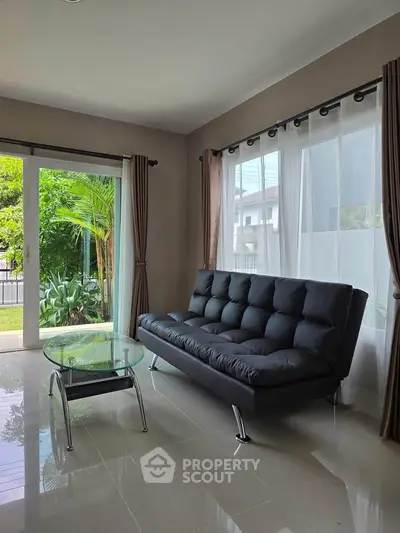 Modern living room with sleek black sofa and glass coffee table, overlooking lush garden through large windows.