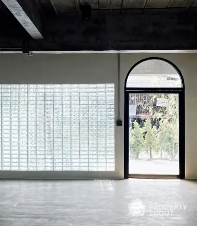 Modern industrial space with glass block wall and arched doorway leading to garden view.