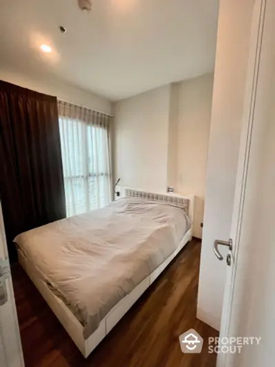 Cozy bedroom with hardwood flooring, large window with curtains, and a comfortable double bed, ideal for restful nights in a modern apartment.