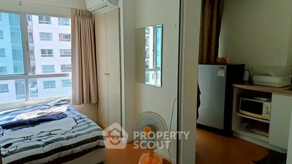 Cozy bedroom with large window, modern furnishings, and compact kitchen area in urban apartment.