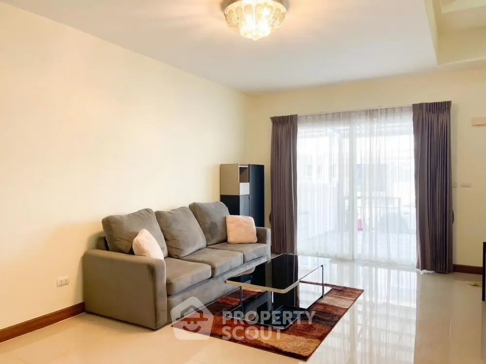 Spacious and well-lit living room with elegant furnishings, large windows draped with maroon curtains, and a cozy grey sofa set on a warm-toned rug.