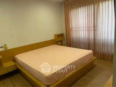 Cozy bedroom with wooden flooring and large window draped with curtains, featuring a comfortable bed.