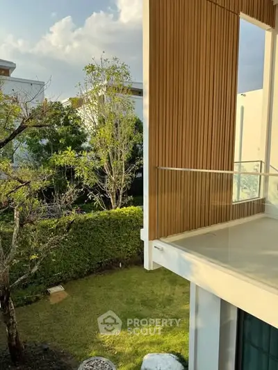 Modern house exterior with glass balcony and lush garden view.