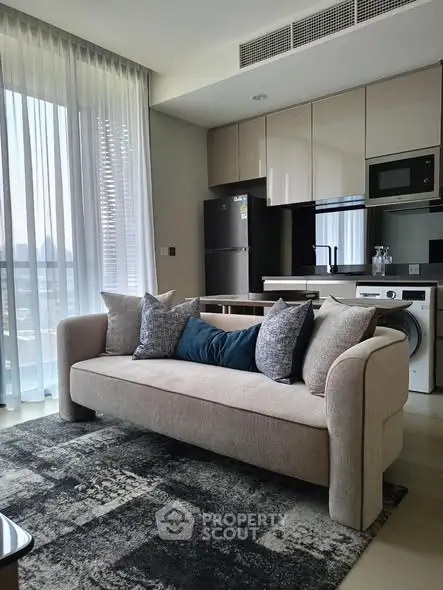 Modern living room with stylish sofa and open kitchen layout, featuring sleek appliances and large windows.